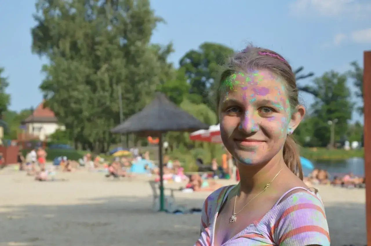 Uśmiechnięta dziewczyna z kolorowym proszkiem na twarzy na plaży zalew Małomice. W tle ludzie odpoczywają na piasku.
