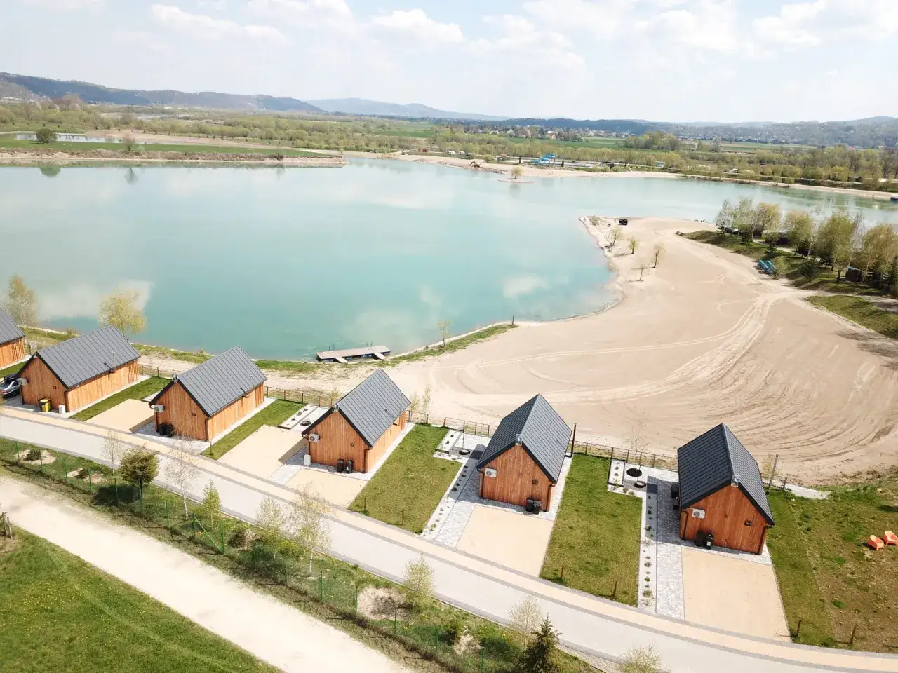 Jurków Mała Chorwacja: drewniane domki nad turkusowym jeziorem, piaszczysta plaża i zielone wzgórza w tle.