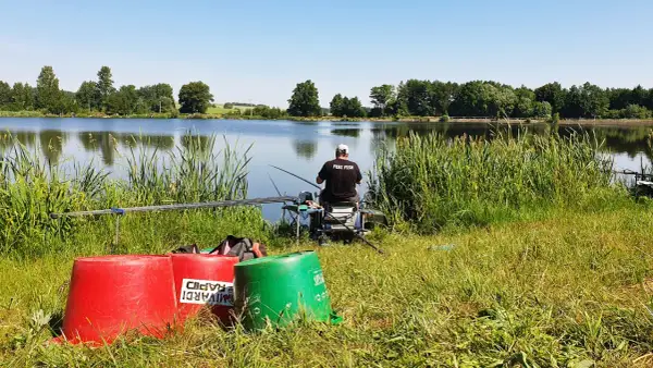 Wędkarz z napisem "FEBI FISH" na koszulce siedzi nad spokojnym stawem, gotowy do łowienia.
