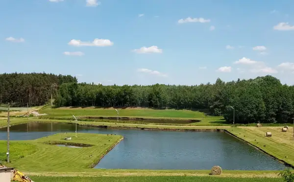 Malownicze łowisko złota rybka z kilkoma stawami, otoczone zielenią i lasem. Na trawiastych brzegach widać beli siana.