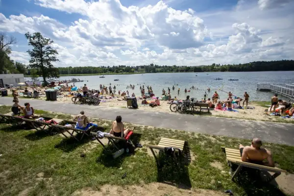 Tłum ludzi odpoczywa na plaży nad jeziorem Kierskim Małym. Słońce świeci, a chmury tworzą malowniczy krajobraz.