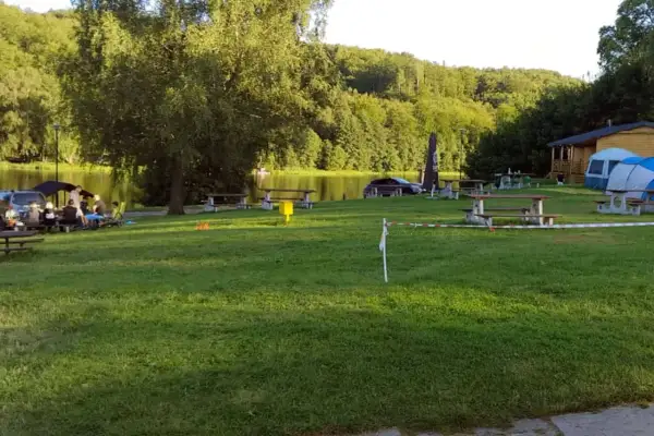 Relaksacyjny camping nad stawami. Ludzie siedzą przy stołach, w tle drzewa i woda. W oddali widać domek i namioty.